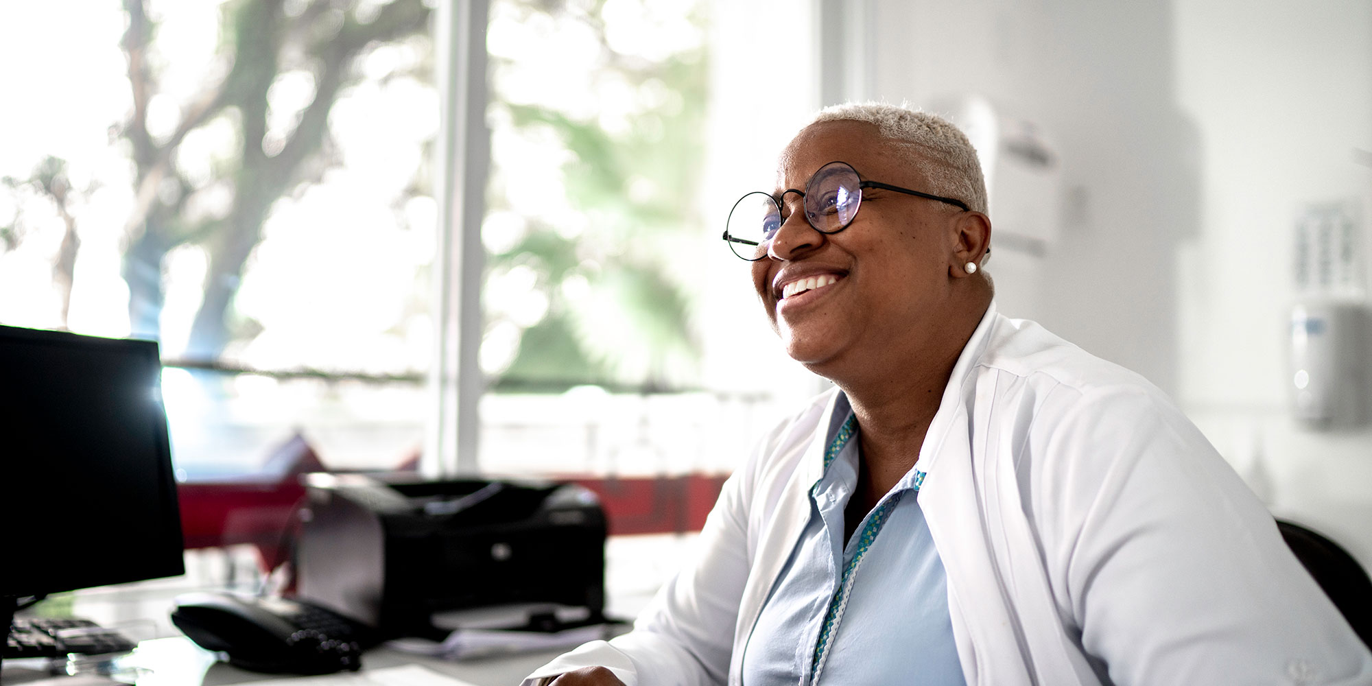 A smiling doctor sits at her desk as she works on paperwork